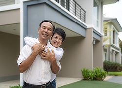 An older couple standing outside their home, hugging and smiling because of their new teeth