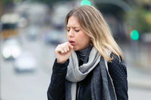 A young woman wearing a jacket and scarf outside while coughing into her hand